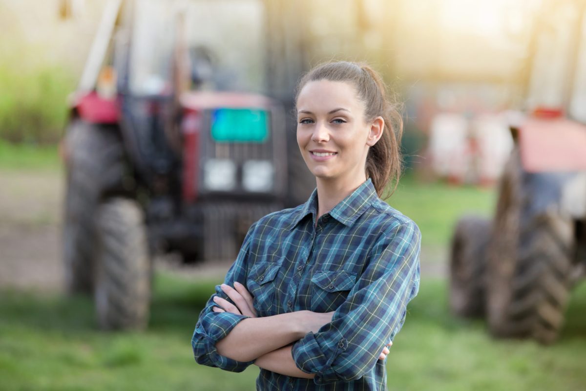 Young pretty farmer woman standing on farmland with crossed arms and tractors in background